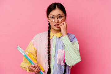Young chinese student woman holding books wearing a fashion multicolour shirt and braid, isolated on pink background biting fingernails, nervous and very anxious.