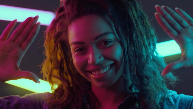 Young happy african american woman looking through her palms and smiling to camera, neon lamps on background
