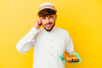 Young Moroccan man wearing the typical arabic costume eating Arabian sweets isolated on yellow background covering ears with hands.