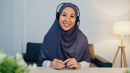 Asia muslim lady wear headphone using laptop talk to colleagues about sale report in conference video call while working from home office at night. Social distancing, quarantine for corona virus.