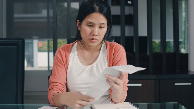 Young Asian Pregnant woman records of income and expenses at home. Mom worried, serious, stress while record budget, tax, financial document working in living room at home concept.