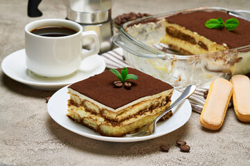 Traditional Italian Tiramisu dessert in glass baking dish and portion on grey concrete background