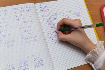 A girls hand with a fountain pen solves examples in mathematics close-up. The student completes the task in teradi. A schoolboy teaches lessons at a school desk. Warm soft daylight.