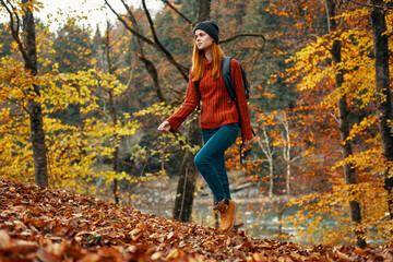 happy woman hiker with a backpack on her back in jeans and a red sweater in the autumn forest park landscape