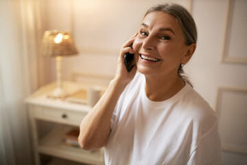 Always in touch. Positive female pensioner posing indoors in casual t-shirt making phone calls, talking to son, glad to hear from him, smiling joyfully at camera. People, age and technology