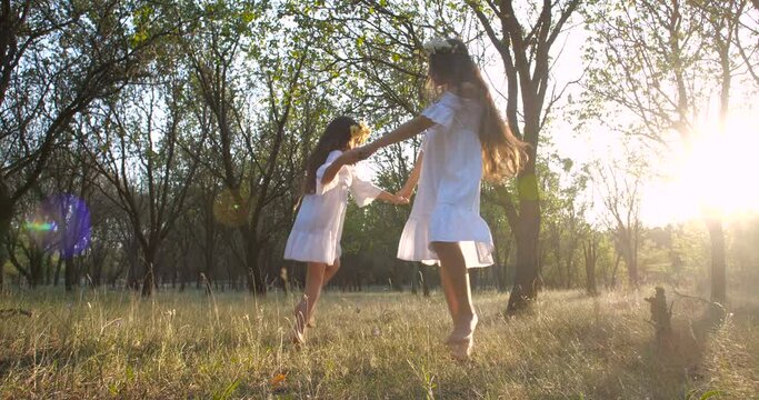 Two little Caucasian sisters with long brown hair  in flower wreaths dancing holding hands on nature background in spring forest. 4k 50 fps slow motion