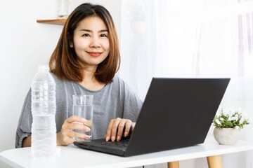 Asian business woman drinking fresh water sitting at office desk hand working on laptop 