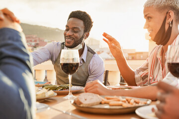 Multiracial people enjoy dinner together outdoor on patio while wearing safety mask under chin - Food and friendship concept