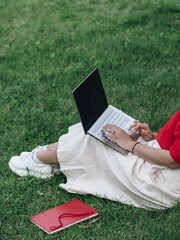 A girl in a red shirt and white skirt is sitting on the grass with a laptop. Study in the fresh air. Remote work in nature. Working on a laptop. The girl has light skin. No face visible.