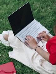 A girl in a red shirt and white skirt is sitting on the grass with a laptop and typing. Study in the fresh air. Remote work in nature. Working on a laptop. The girl has light skin. No face visible.