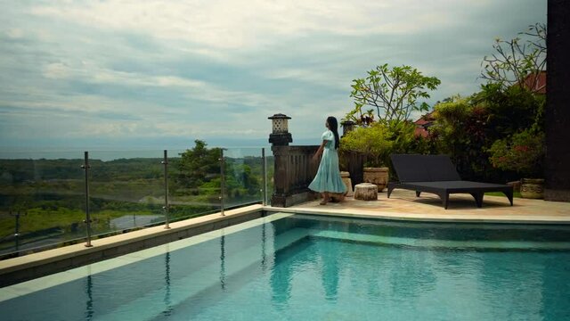 girl walking in the backyard with a green garden, pool with blue clean water and blue sky in a beautiful long blue dress