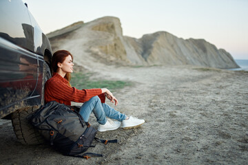 a traveler sits near a car in the mountains in nature and admires the landscape at sunset