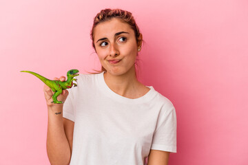Young blonde caucasian woman holding a dinosaur miniature isolated on pink background confused,...