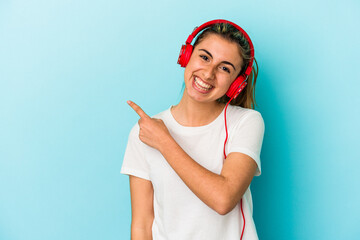 Young blonde woman listening to music on headphones isolated on blue background smiling and pointing aside, showing something at blank space.