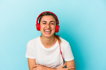 Young blonde woman listening to music on headphones isolated on blue background laughing and having fun.