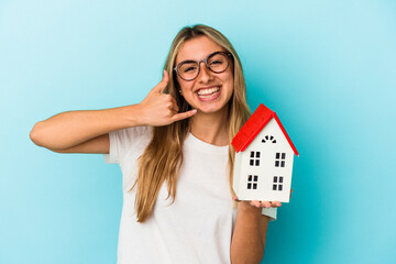 Young caucasian woman holding a house model isolated on blue background showing a mobile phone call...