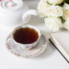 A porcelain tea cup with a saucer, a teapot, a white faience angel and three roses on the table. Breakfast.