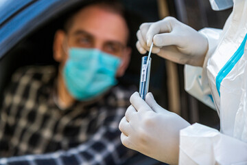 Man sitting in car, waiting for medical worker in PPE to perform drive-thru COVID-19 test
