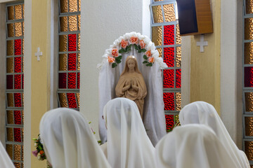 Veiled heads of nuns praying and singing before an image of the Virgin Mary in a flowered niche in...