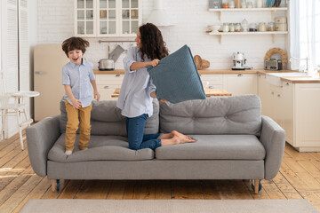 Mom and son have fun in living room fight with pillows together. Family leisure and recreation time. Cute cheerful and joyful young mother and little boy laugh while playing funny battle games at home