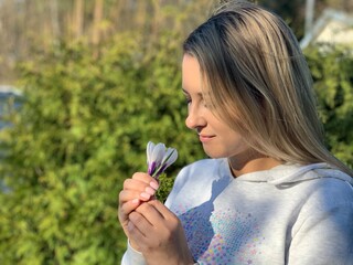 A beautiful blonde girl holding white crocus flowers in her hand, smiling and sniffing at them against the background of green tree branches.