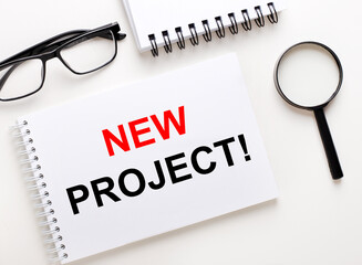 NEW PROJECT is written in a white notebook on a light background near the notebook, black-framed glasses and a magnifying glass.