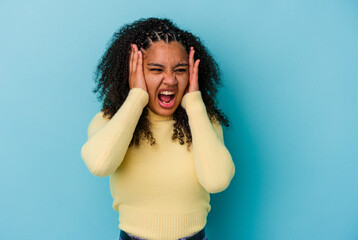 Young african american woman isolated on blue background covering ears with hands trying not to hear too loud sound.