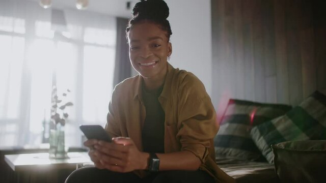 Young african american attractive woman smiling on camera using her mobile phone social media application shopping online from home. Portraits. Interiors.