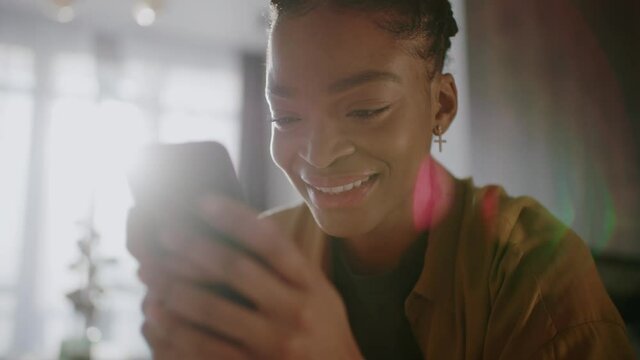 Face of beautiful modern young adult african american woman holding mobile phone typing messages on social media. Home interior background.