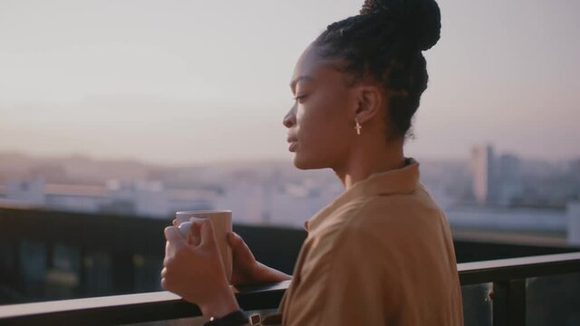 Young inspired woman standing on her terrace, enjoying drinking tea, watching views of sunrise landscape and scenery. People at home. Peaceful morning.