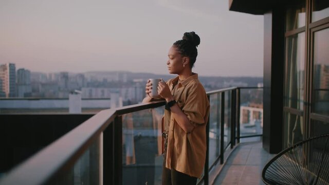 Lovely african american woman in casual outfit walking on her terrace balcony, drinking coffee in cup and looking at sunrise.