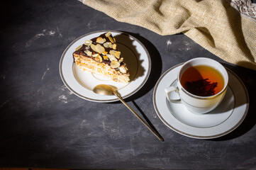 Still life - layered homemade sponge cake and a cup of black tea in a white dish, and a linen napkin, black background, hard light, photo in a low key.