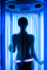 Young sexy female in a tanning cabin of vertical solarium. girl sunbathing in solarium cabin with blue luminous tanning lamps in bikini - obrazy, fototapety, plakaty
