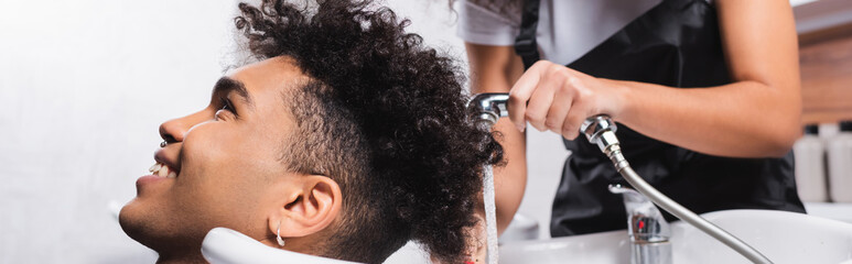 Smiling african american client sitting near hairdresser with shower on blurred background, banner