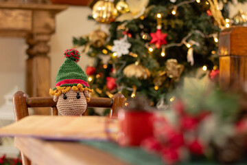 Christmas time. Selective focus on Santa's helper, the crochet elf sitting in the living room reading. Festive xmas tree decorated with warm lights on blurry background.