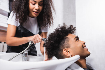 Positive african american client sitting near hairdresser with shower on blurred background in salon