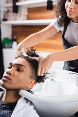 Obraz na płótnie Canvas African american hairdresser washing hair of young man near sink