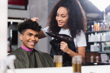 Smiling client looking at mirror near hairstylist with hair dryer in salon