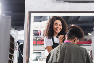 African american hairdresser smiling at camera while holding trimmer near man in salon