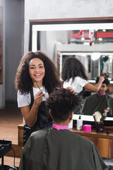 Positive african american hairdresser with comb and scissors working with client in salon