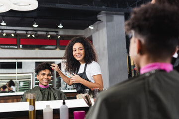 Smiling hairstylist holding comb near hair of african american client on blurred foreground