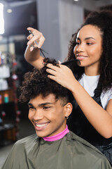 Smiling african american client in cape sitting near hairdresser cutting hair on blurred background