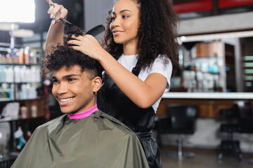 Cheerful african american client sitting near hairdresser holding scissors and comb in salon