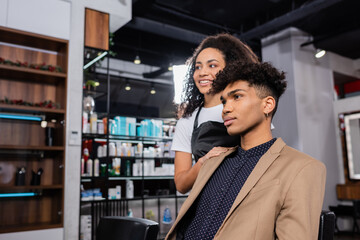 Positive african american hairstylist on blurred background standing near young man