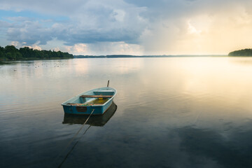 Wooden boat floating on the shore of a lake in Elk in the Mazury province of Poland. A storm comes in the background, as the sun disappears, calm before the storm. - obrazy, fototapety, plakaty
