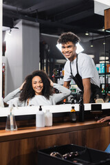Smiling african american hairdresser and client looking at mirror in salon