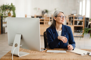 The Asian woman with glasses sitting at a table in front of a computer and looking to the side in...