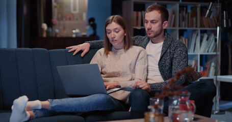 Attractive caucasian couple of cherful woman and man chatting indoors having fun communicating using laptop choosing comedy movie watching at night.