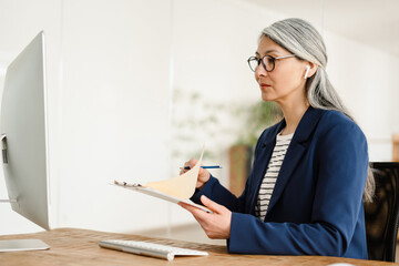 The side view of Asian woman flipping paper on tablet and looking at monitor camera in office