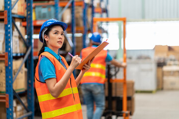 Asian young female employee warehouse worker with safety vest and helmet using clipboard for...